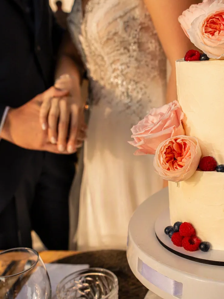Tarta de boda de dos pisos en crema con rosas inglesas rosadas y frutos rojos, junto a los novios cogidos de la mano, en un venue de Granada — Magda Sweet Cakes.